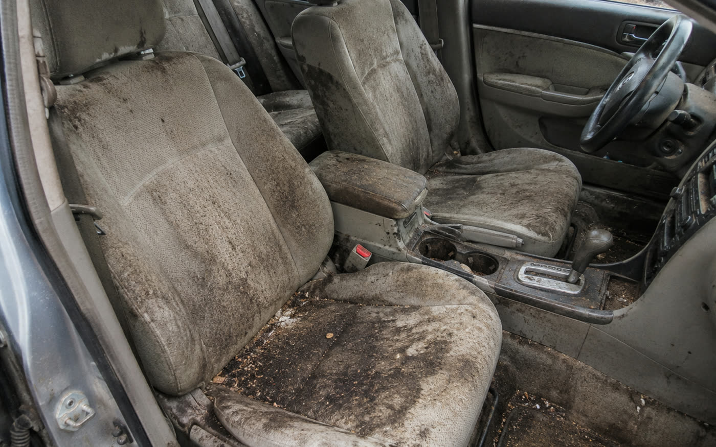 The front seats and centre console of a car, the pale cloth upholstery and carpet darkened and matted with ground-in dirt